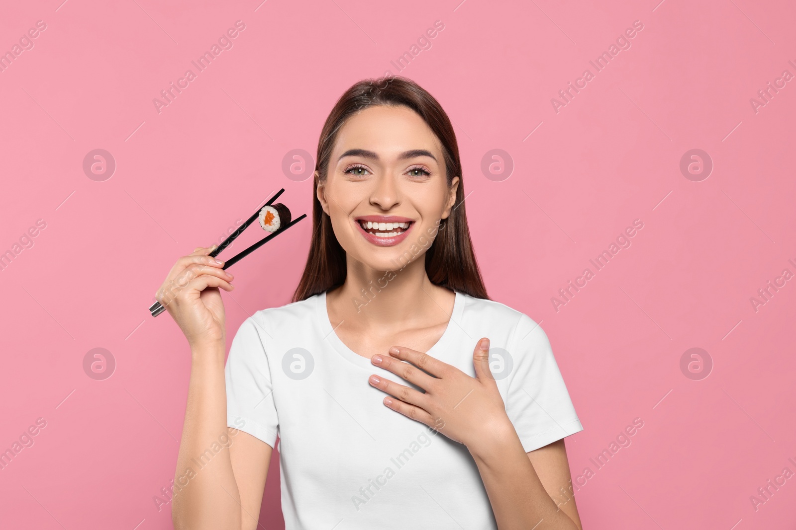 Photo of Beautiful young woman holding sushi roll with chopsticks on pink background