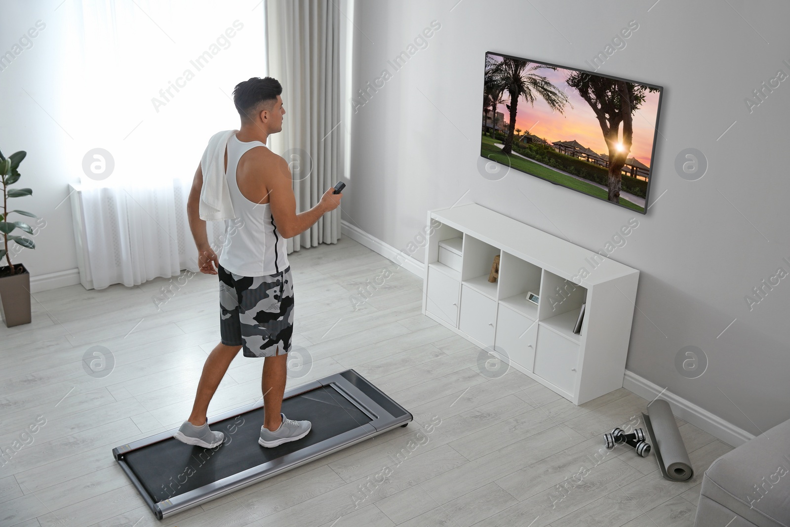 Image of Sporty man with remote control training on walking treadmill and watching TV at home