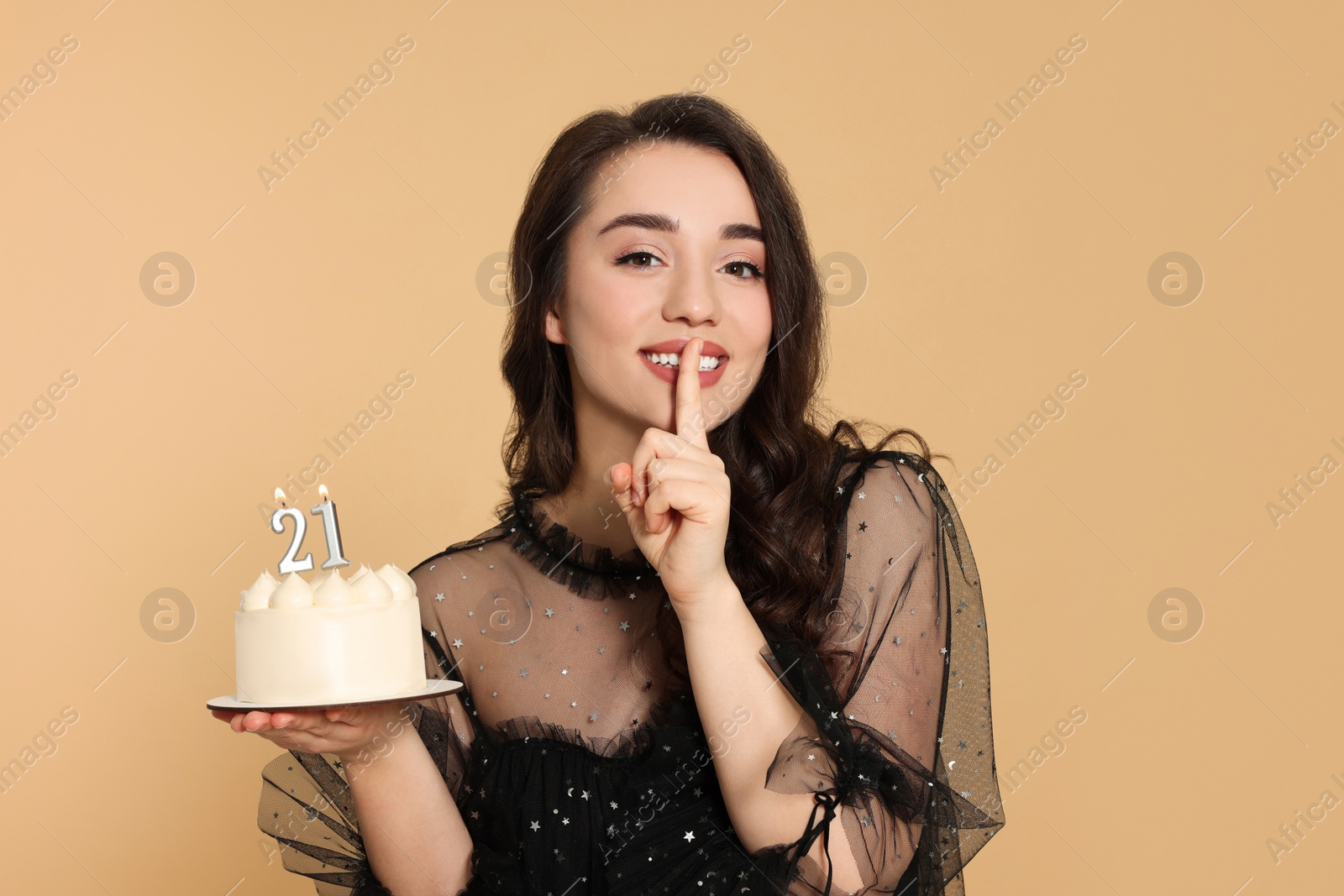 Photo of Coming of age party - 21st birthday. Smiling woman showing silence gesture and holding delicious cake with number shaped candles on beige background