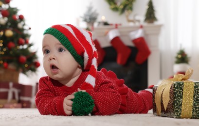 Photo of Baby in elf hat near Christmas gift on floor at home