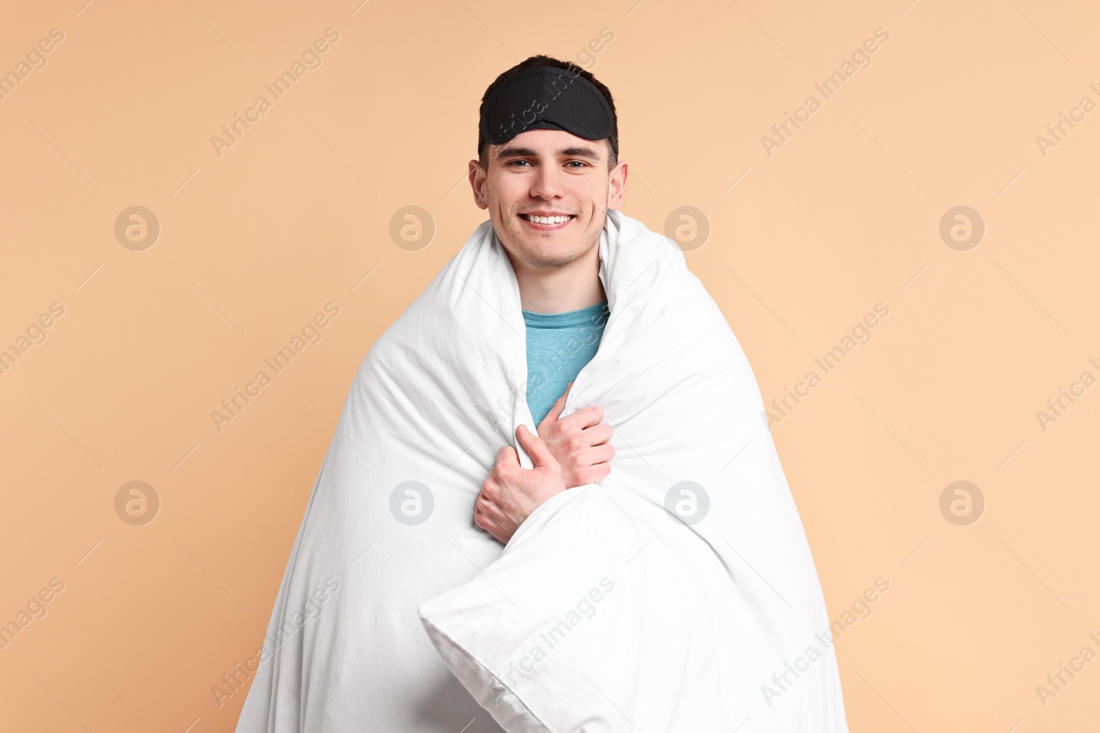 Photo of Happy man in pyjama and sleep mask wrapped in blanket on beige background