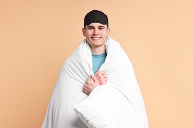 Happy man in pyjama and sleep mask wrapped in blanket on beige background