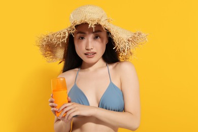Beautiful young woman in straw hat holding sun protection cream on orange background