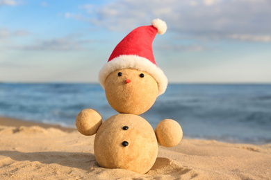 Snowman made of sand with Santa hat on beach near sea. Christmas vacation