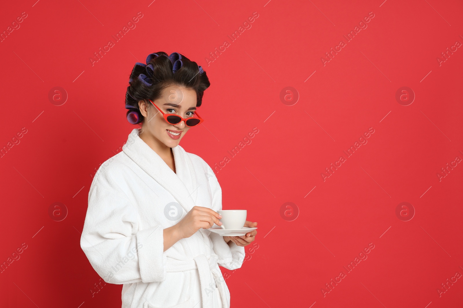 Photo of Happy young woman in bathrobe with hair curlers and sunglasses holding cup of drink on red background, space for text