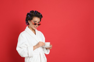 Happy young woman in bathrobe with hair curlers and sunglasses holding cup of drink on red background, space for text