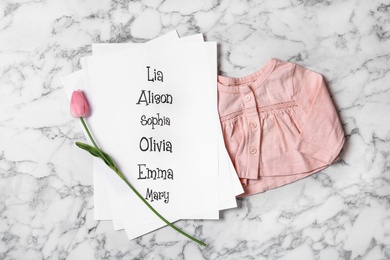 List of baby names, tulip flower and child's clothes on white marble background, flat lay
