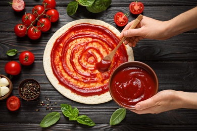 Photo of Woman spreading tomato sauce onto pizza crust on dark wooden table, top view