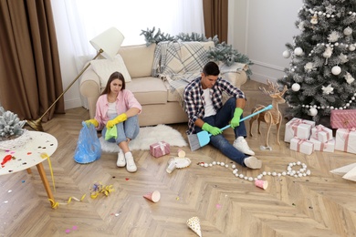 Photo of Tired couple sitting in messy room while cleaning after New Year party