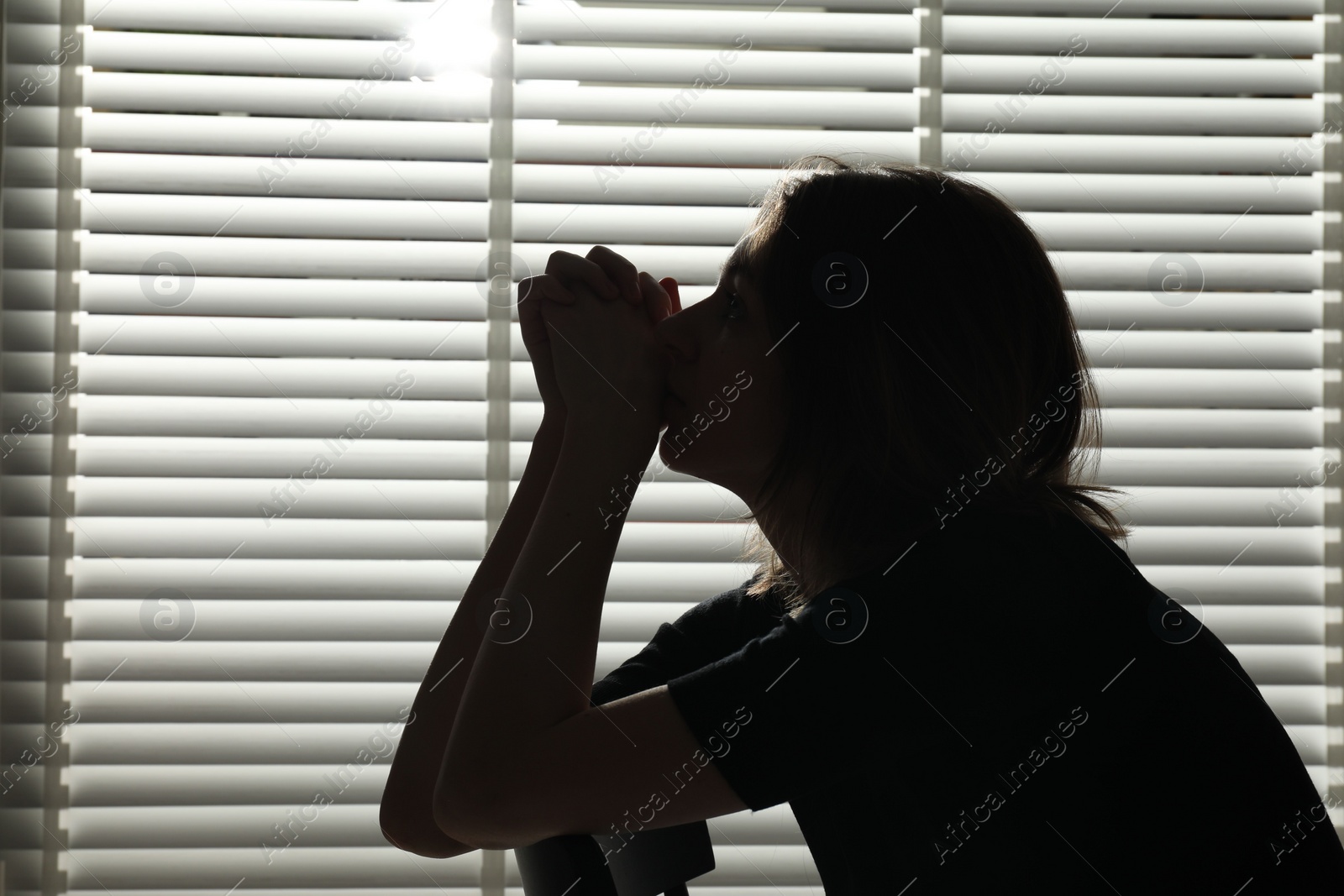 Photo of Silhouette of sad young woman near closed blinds indoors, space for text