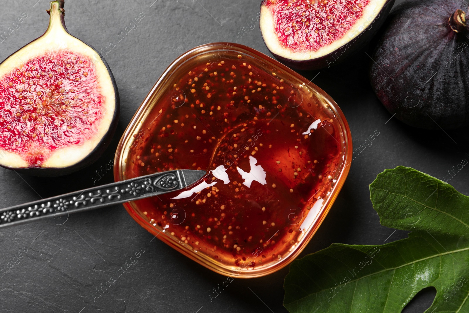 Photo of Glass bowl with tasty sweet jam, spoon, fresh figs and green leaf on grey table, flat lay