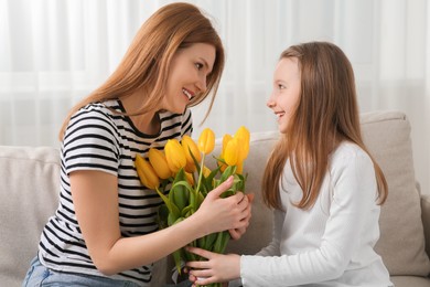 Happy mother and her cute daughter with bouquet of yellow tulips on sofa at home