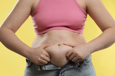 Woman touching belly fat on yellow background, closeup. Overweight problem