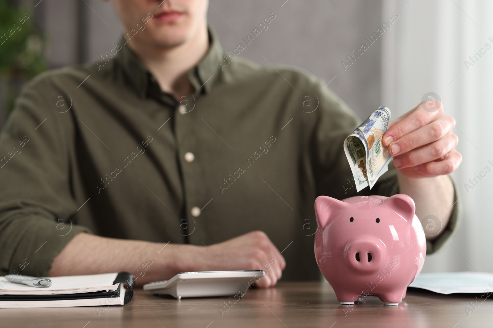 Photo of Financial savings. Man putting dollar banknote into piggy bank at wooden table, closeup