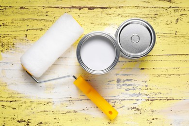 Photo of Can of white paint and roller on yellow wooden table, flat lay