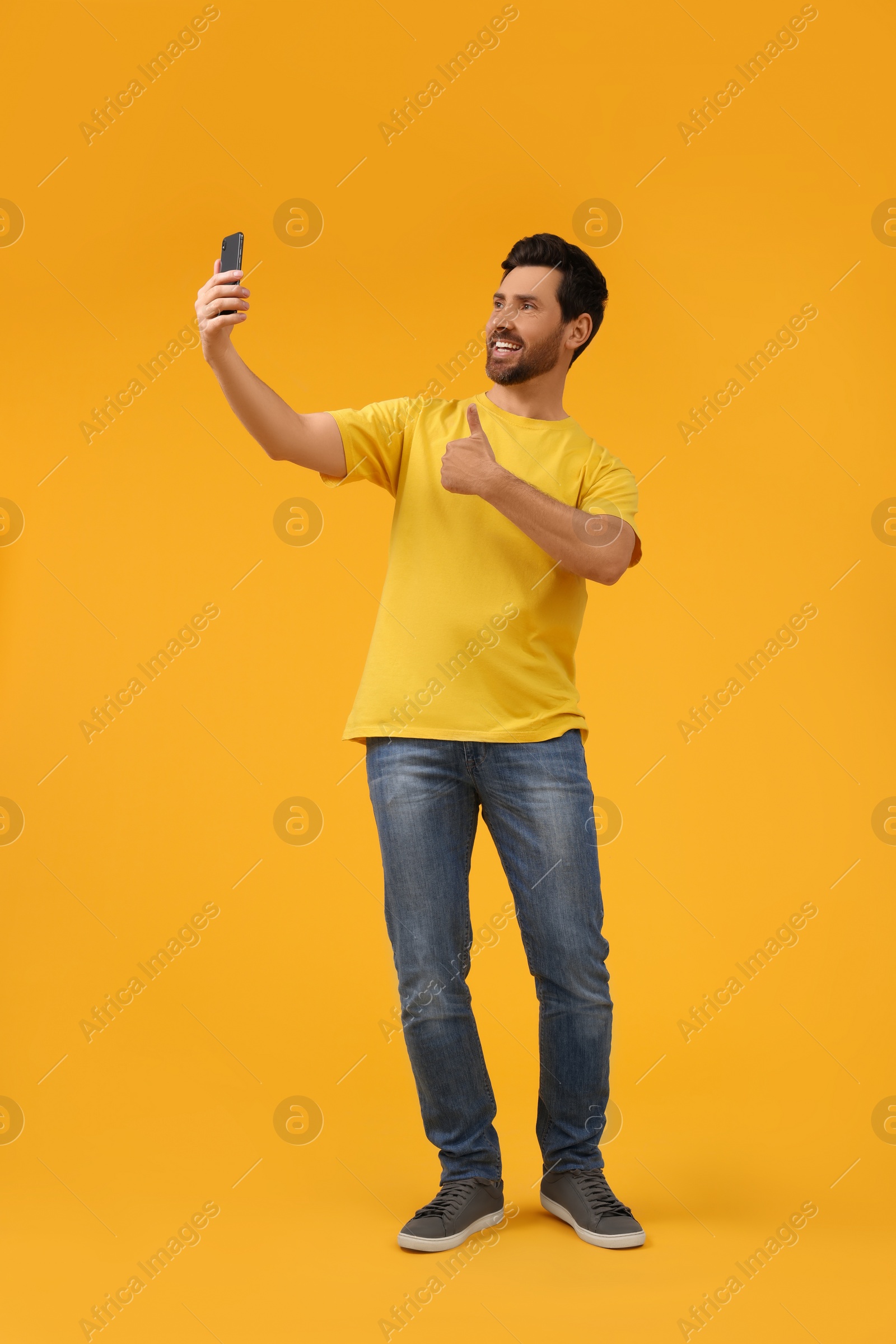 Photo of Smiling man taking selfie with smartphone and showing thumbs up on yellow background, space for text