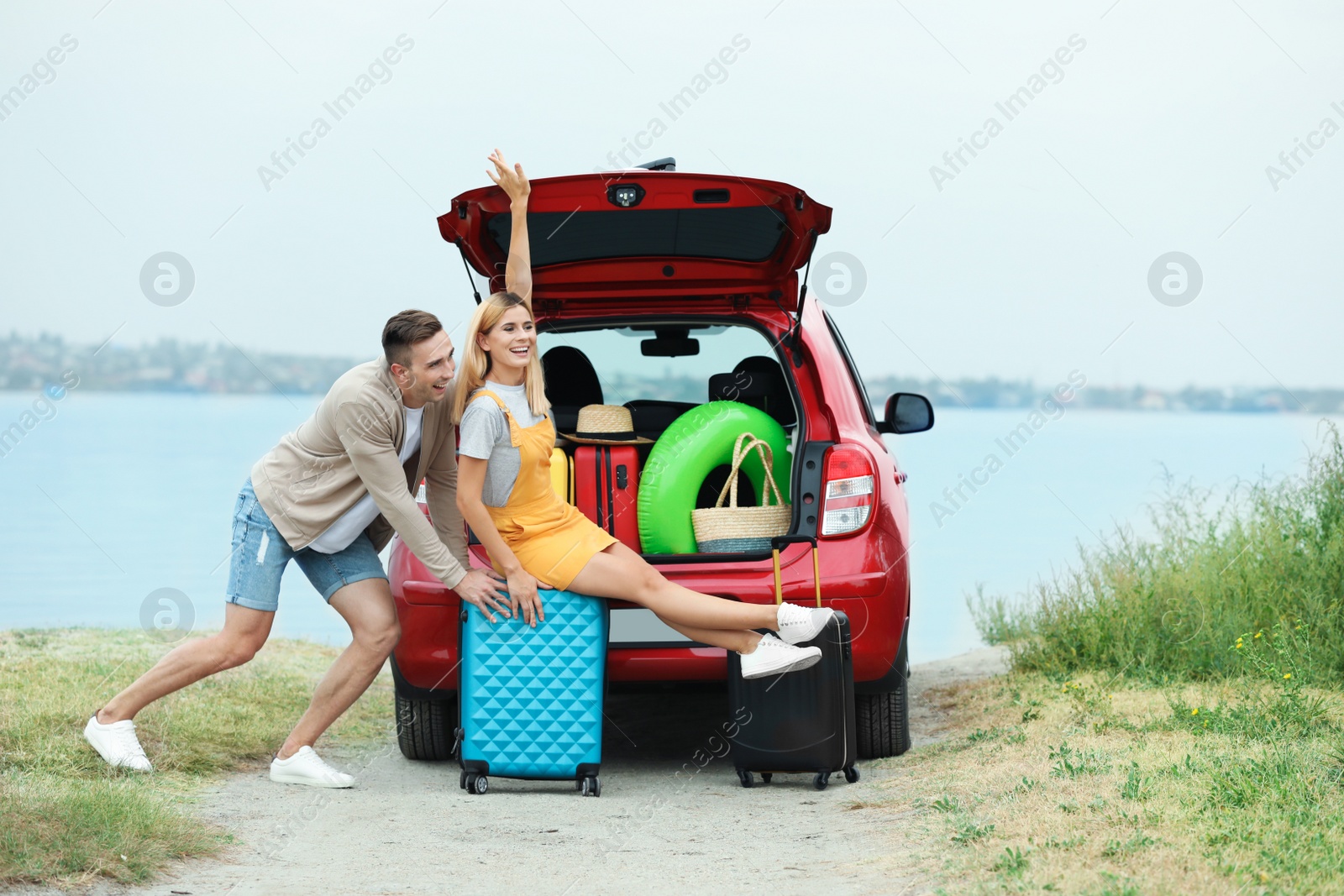 Photo of Couple having fun near car trunk with suitcases on riverside