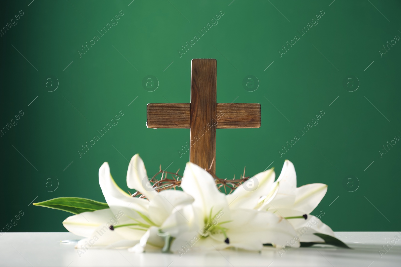 Photo of Wooden cross, crown of thorns and blossom lilies on table against color background