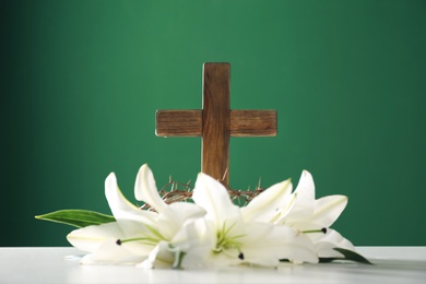 Photo of Wooden cross, crown of thorns and blossom lilies on table against color background