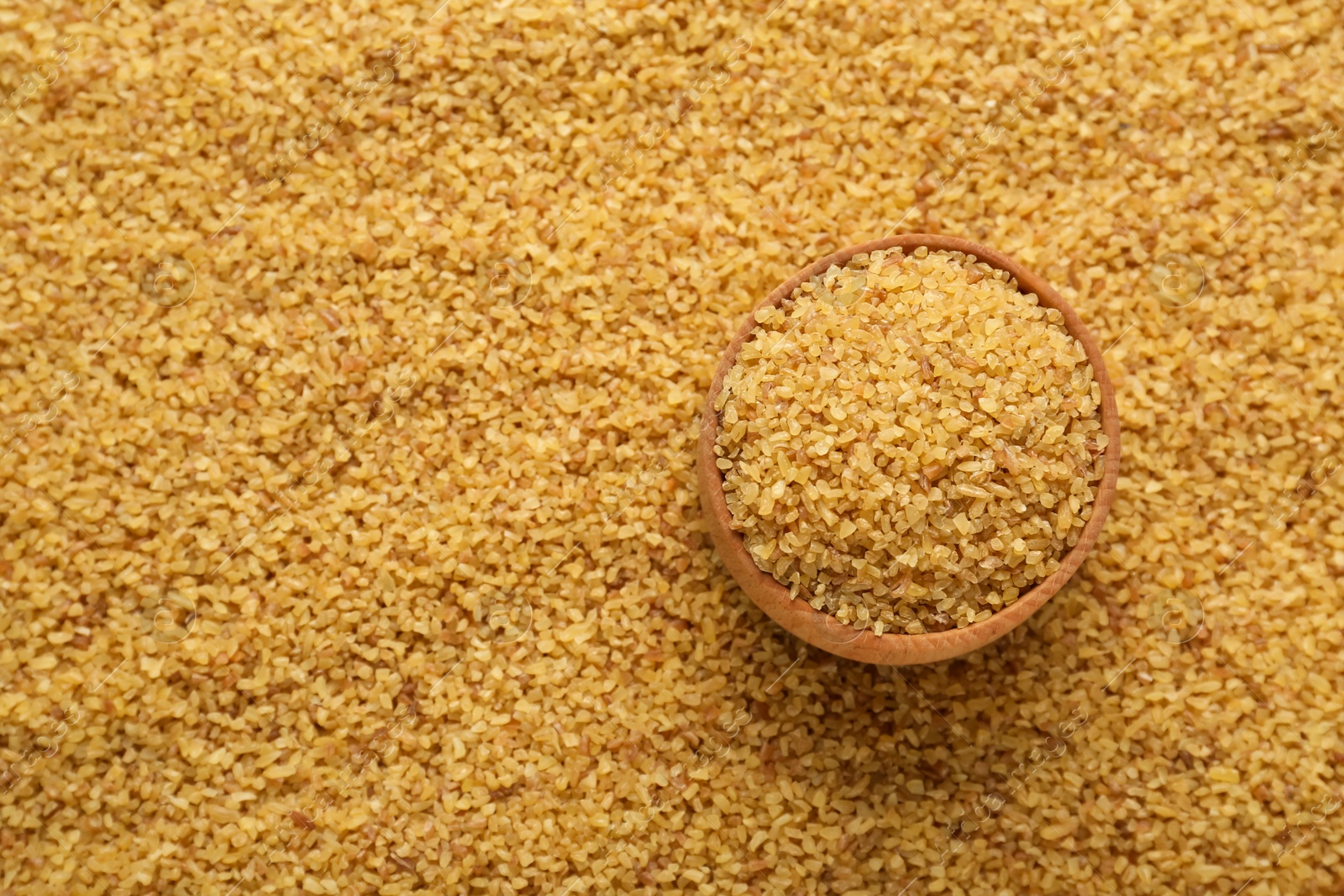 Photo of Wooden bowl on pile of uncooked bulgur, top view. Space for text