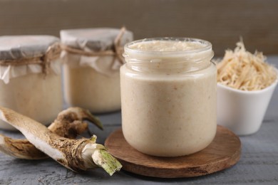 Photo of Spicy horseradish sauce in jar and roots on grey wooden table, closeup