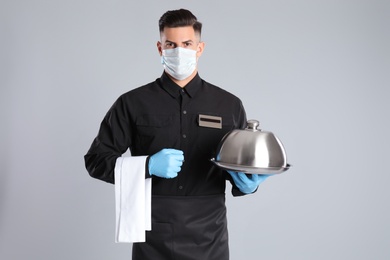 Waiter in medical face mask holding tray with lid on light grey background