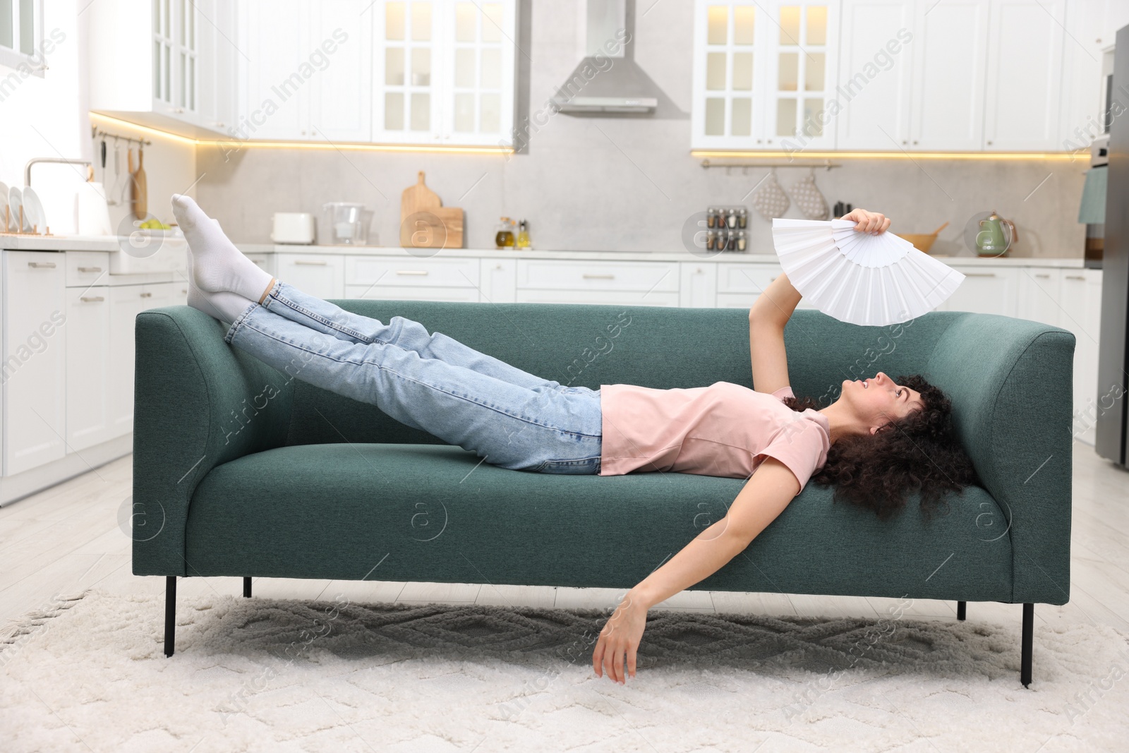 Photo of Young woman waving white hand fan to cool herself on sofa at home