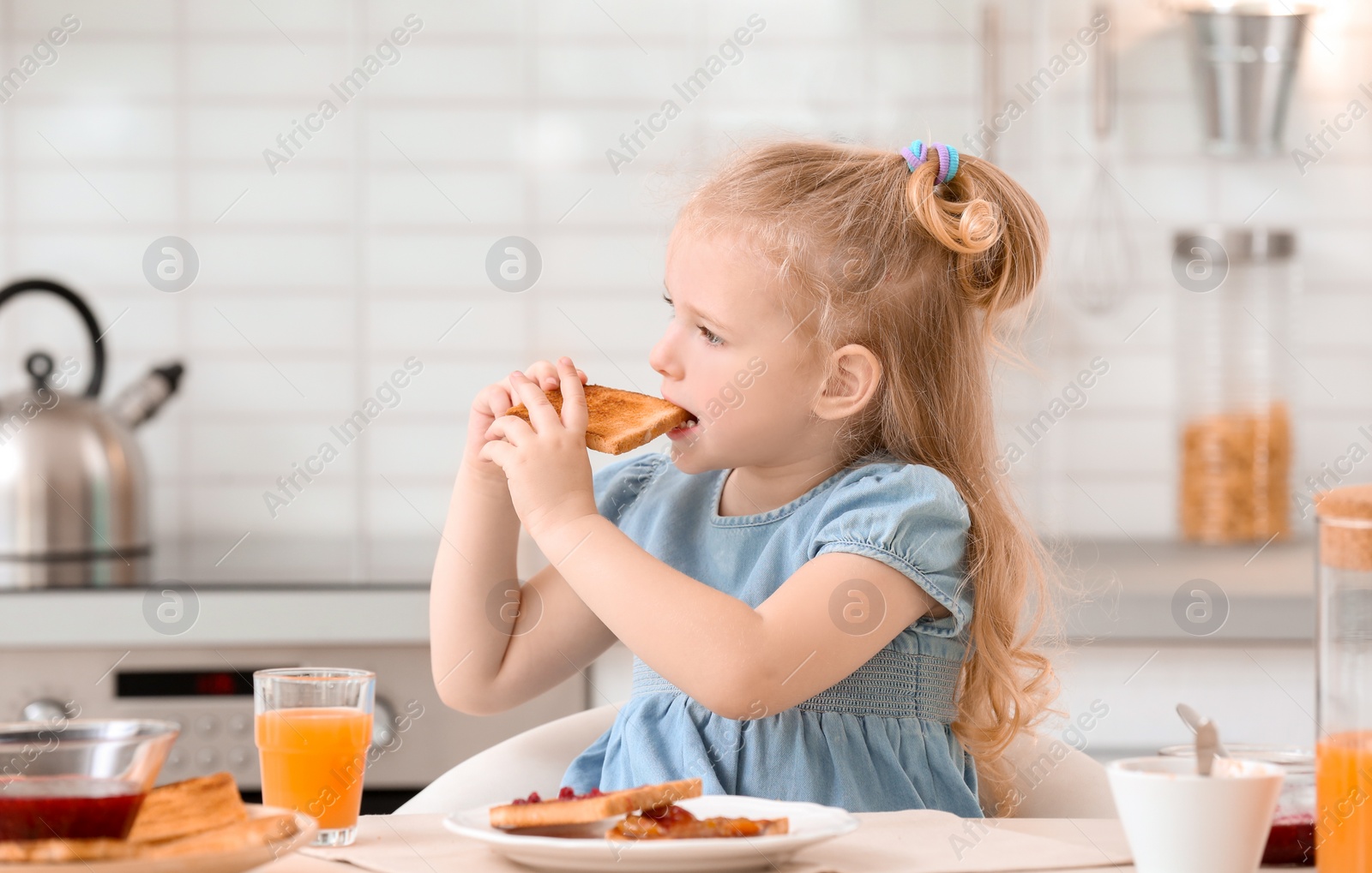 Photo of Adorable little girl eating tasty toasted bread with jam at table in kitchen