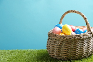Wicker basket with painted Easter eggs on green grass against color background, space for text