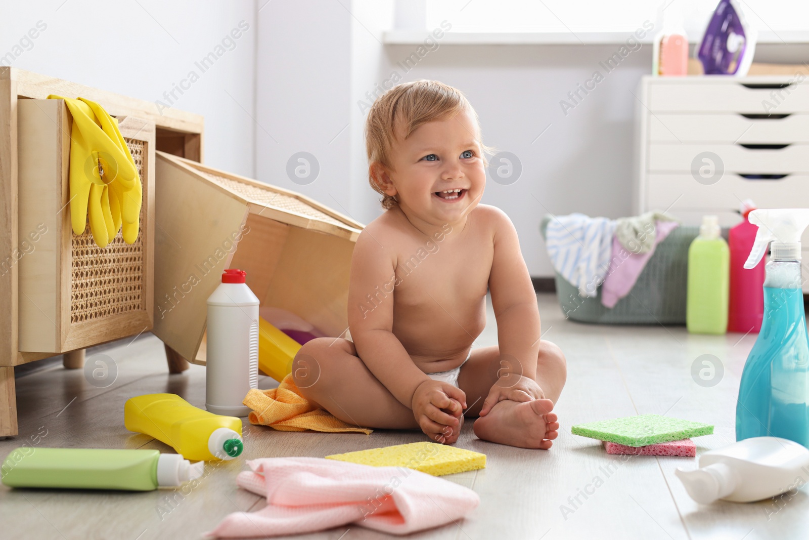 Photo of Cute baby surrounded by cleaning supplies at home. Dangerous situation