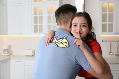 Photo of Cute little girl sticking paper fish to father's back at home