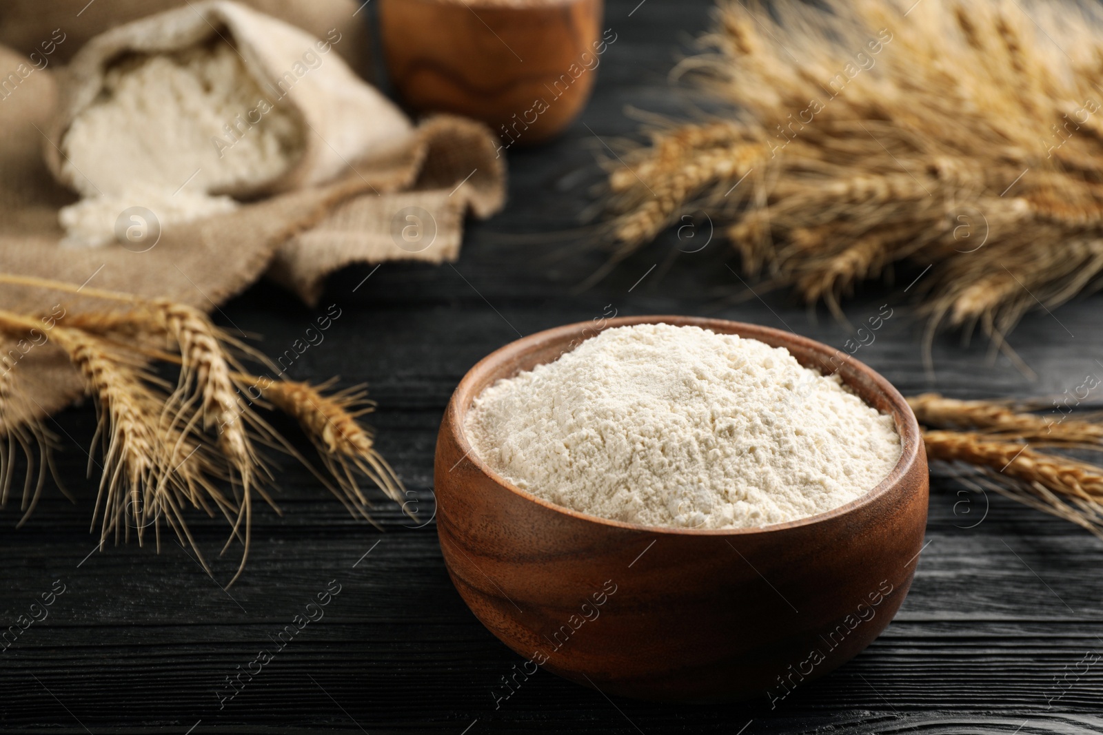 Photo of Bowl of flour and wheat ears on black wooden table