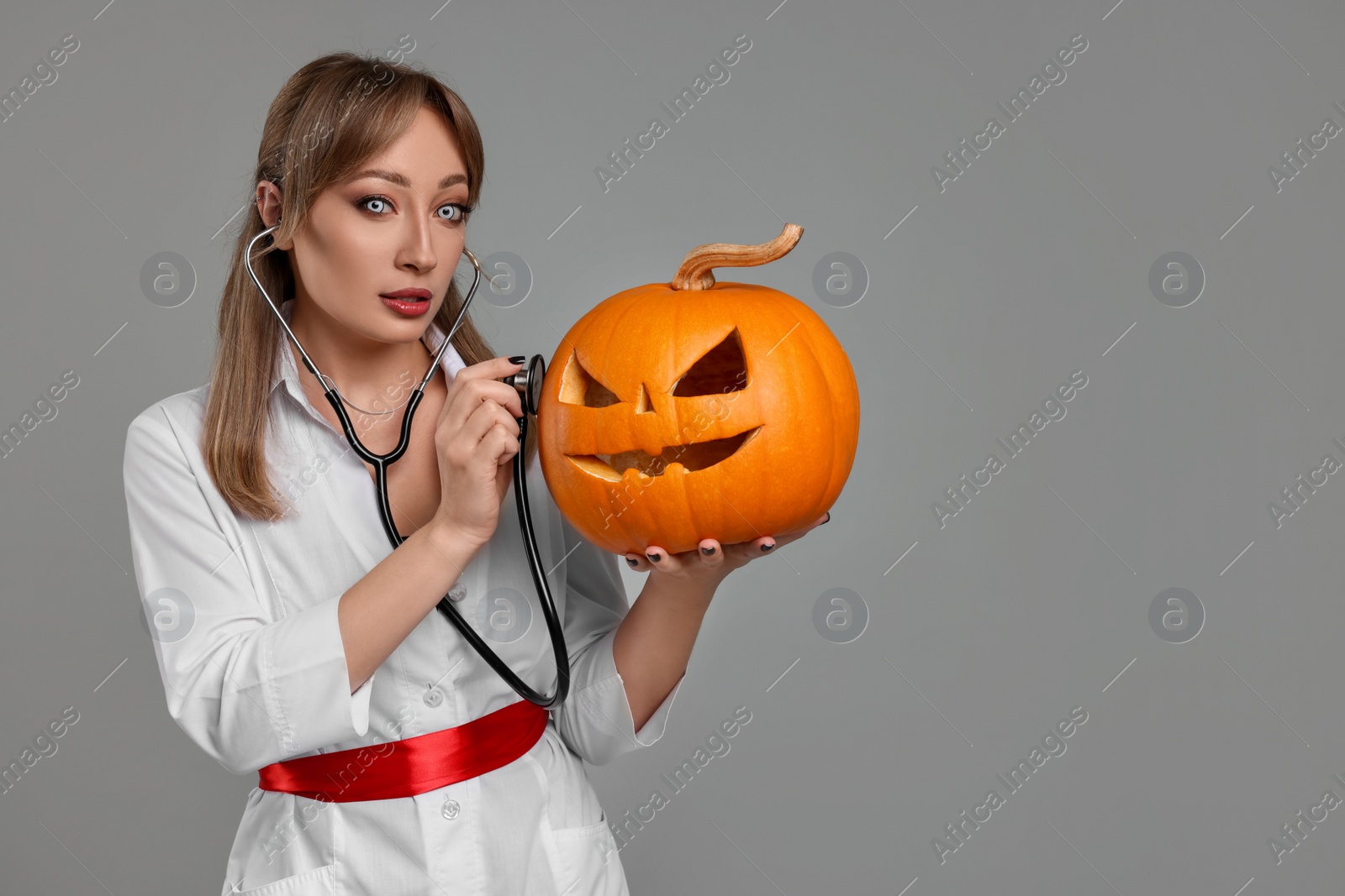 Photo of Woman in scary nurse costume with carved pumpkin and stethoscope on light grey background, space for text. Halloween celebration