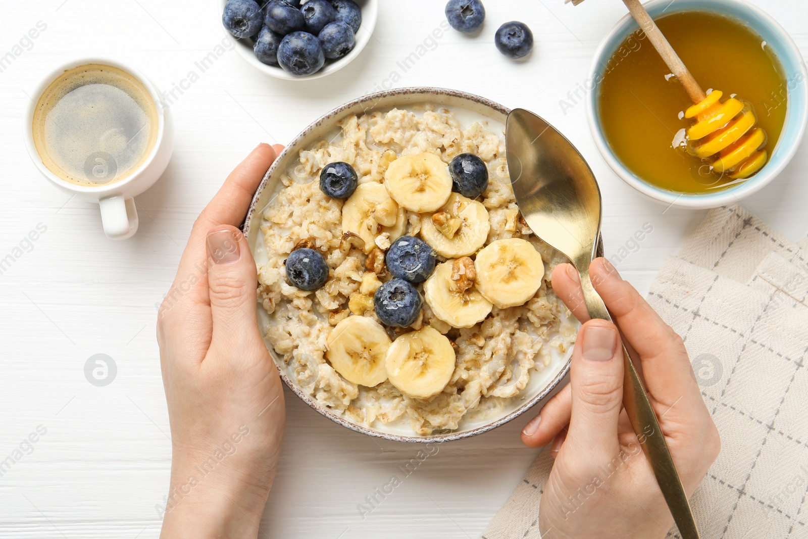 Photo of Woman eating tasty oatmeal with banana, blueberries, walnuts and honey at white wooden table, top view