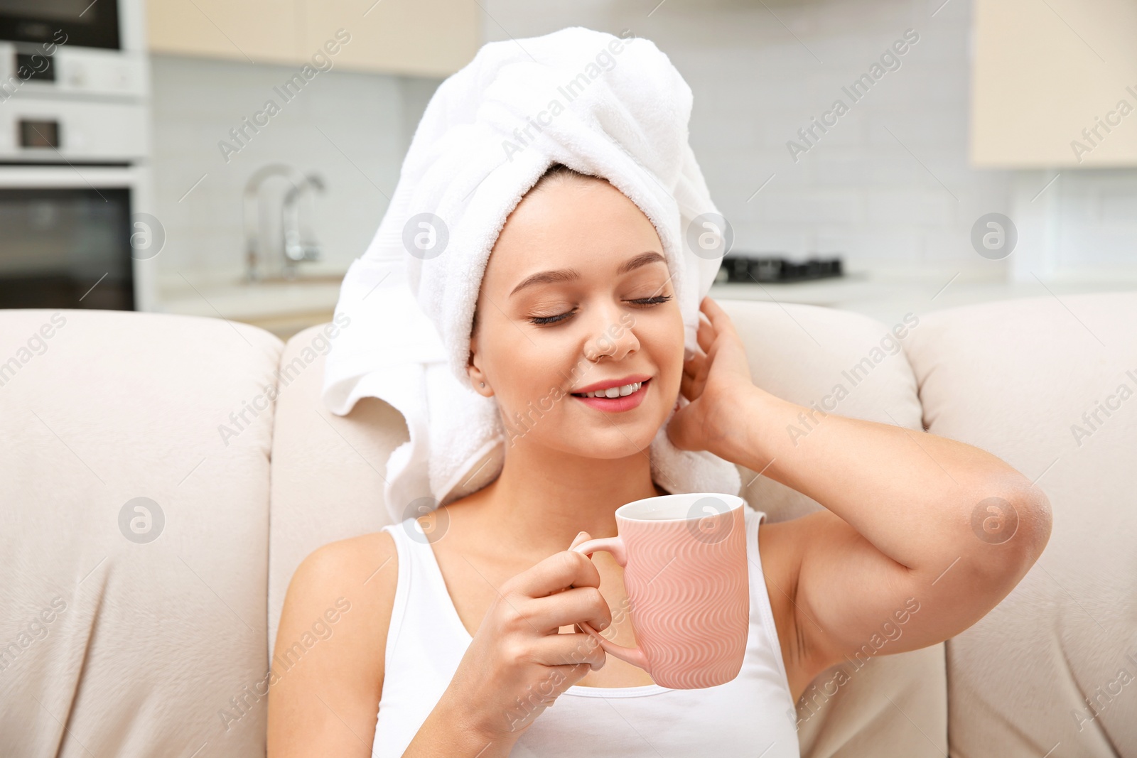 Photo of Beautiful woman with hair wrapped in towel sitting on sofa and drinking tea at home