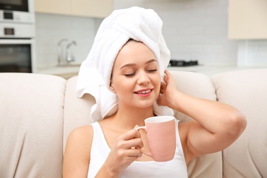 Beautiful woman with hair wrapped in towel sitting on sofa and drinking tea at home
