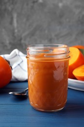 Photo of Delicious persimmon jam in glass jar and fresh fruits on blue wooden table