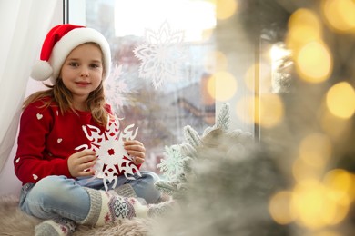 Photo of Little girl in Santa hat with paper snowflake near small Christmas tree at home