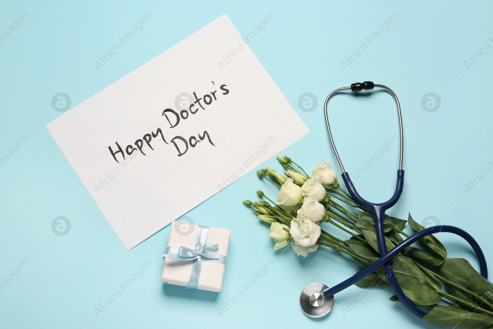 Photo of Card with phrase Happy Doctor's Day, stethoscope, gift box and eustoma flowers on light blue background, flat lay