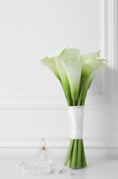Photo of Beautiful calla lily flowers in glass vase, bottle of perfume and jewelry on white table
