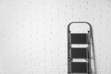 Photo of Modern metal stepladder against white brick background. Space for text