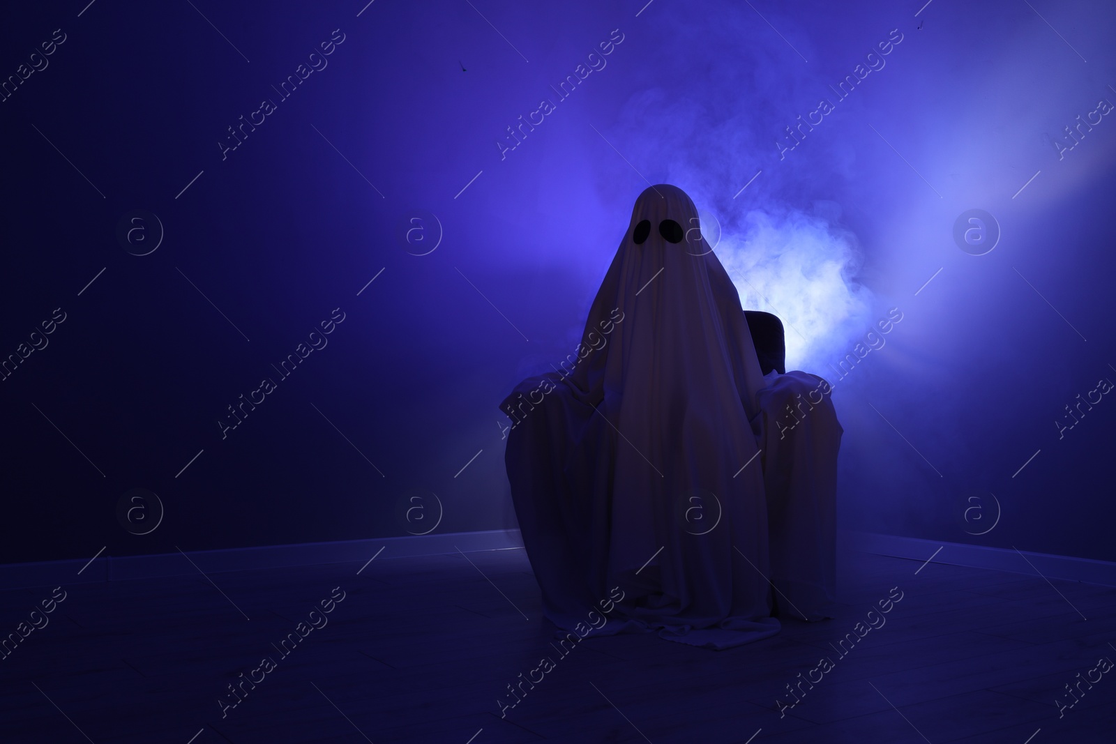 Photo of Creepy ghost. Woman covered with sheet sitting in armchair in blue light, space for text