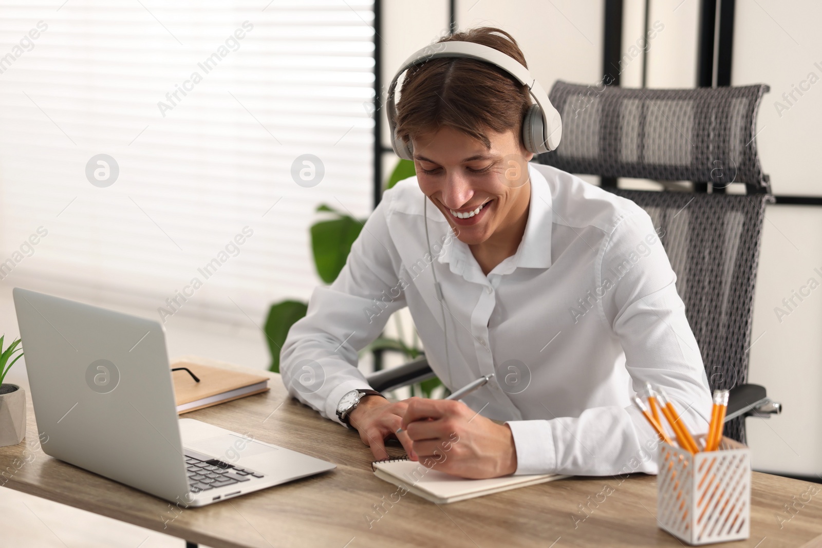 Photo of Man in headphones taking notes during webinar at wooden table indoors
