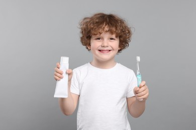 Photo of Cute little boy holding electric toothbrush and tube of toothpaste on light grey background
