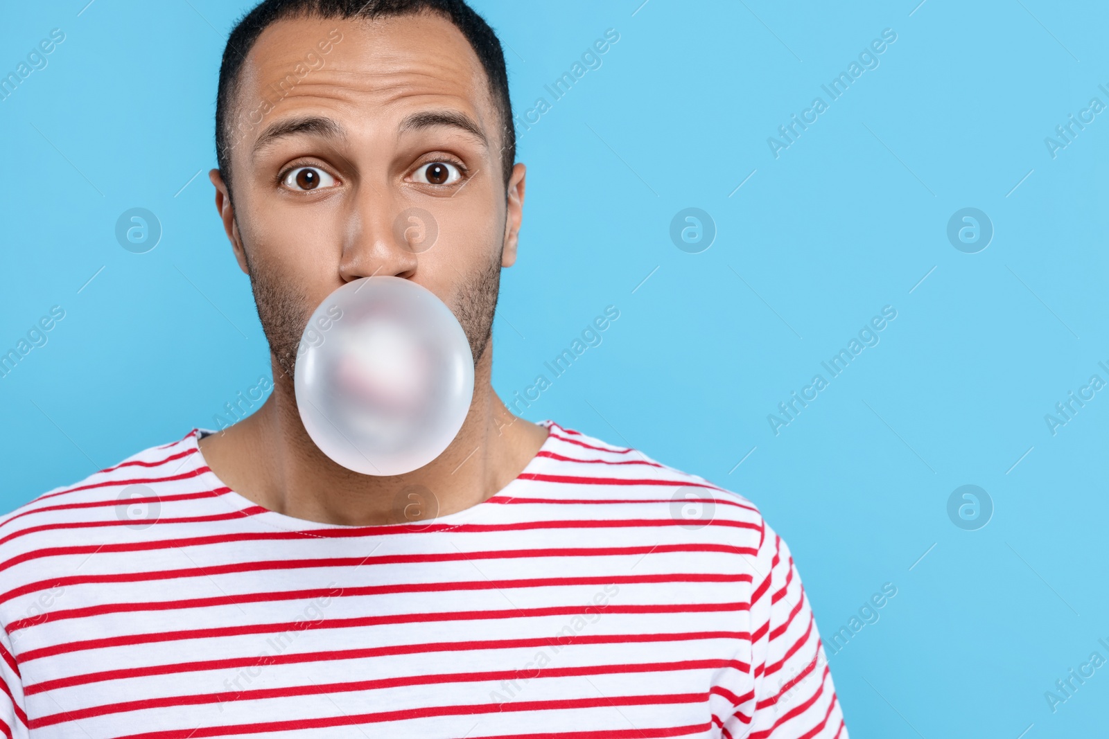 Photo of Portrait of young man blowing bubble gum on light blue background. Space for text