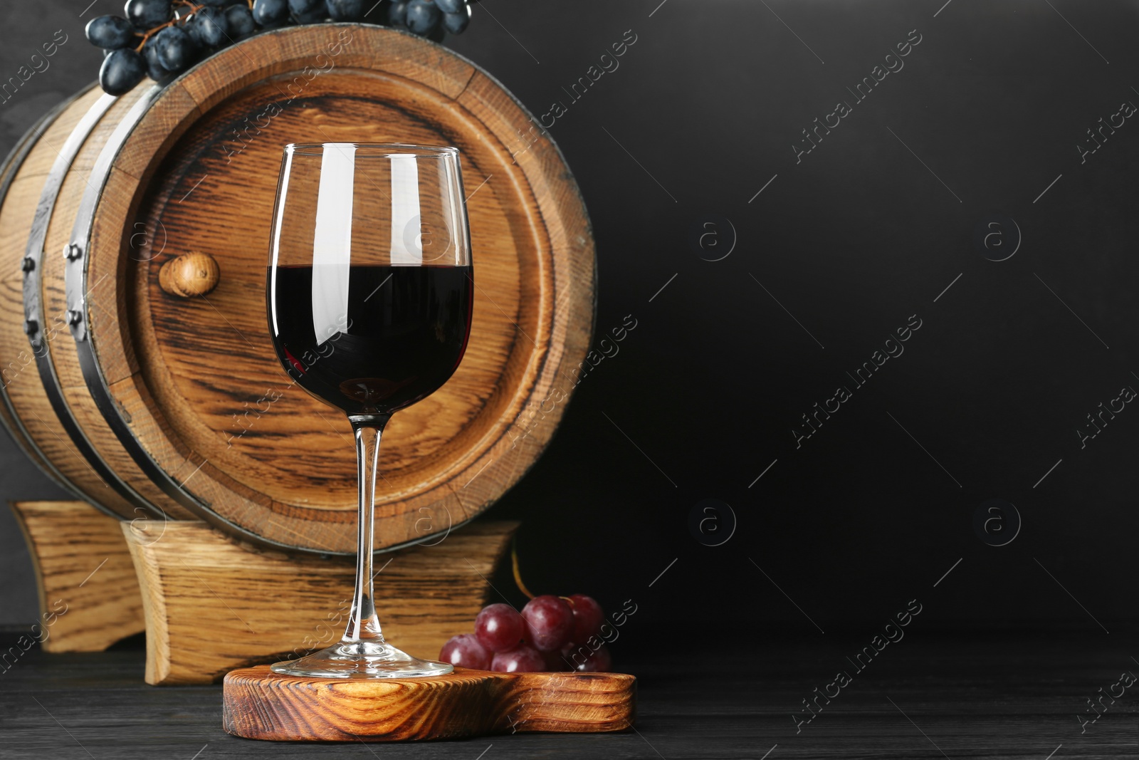 Photo of Glass with delicious wine, wooden barrel and fresh grapes on black table, space for text