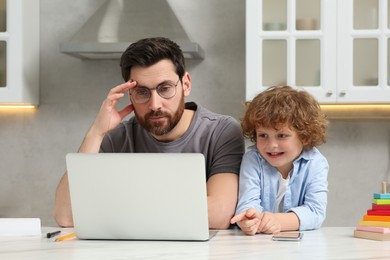 Photo of Little boy bothering father while he working remotely at home. Man with laptop and his child at desk in kitchen