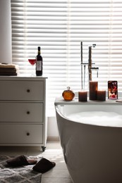 Photo of White wooden tray with smartphone and burning candles on bathtub in bathroom