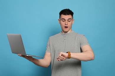 Emotional young man with laptop checking time on light blue background. Being late concept