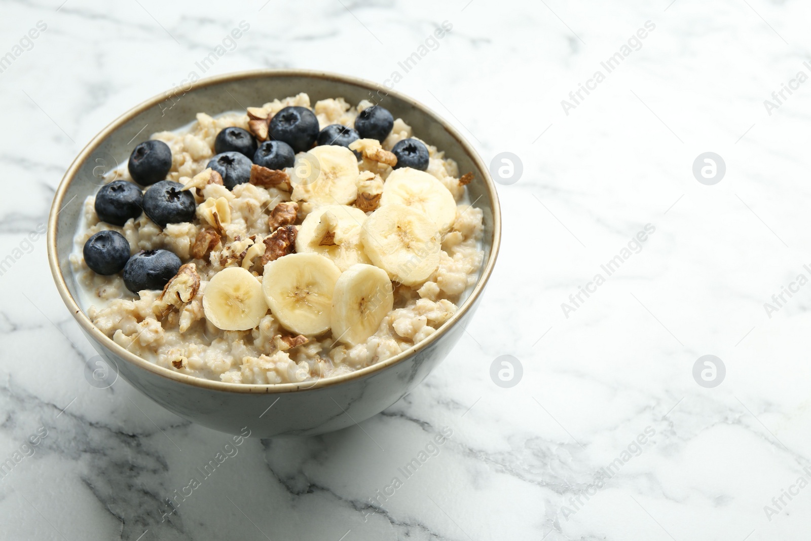 Photo of Tasty oatmeal with banana, blueberries, walnuts and milk served in bowl on white marble table, space for text